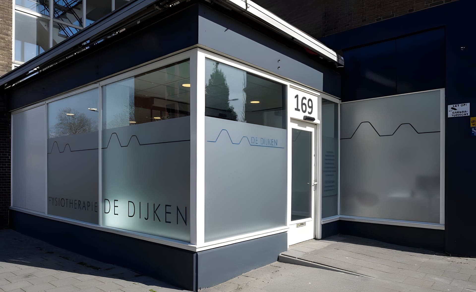 Het hoekpand van Fysiotherapie De Dijken met huisnummer 169 en de naam in de beglazing.