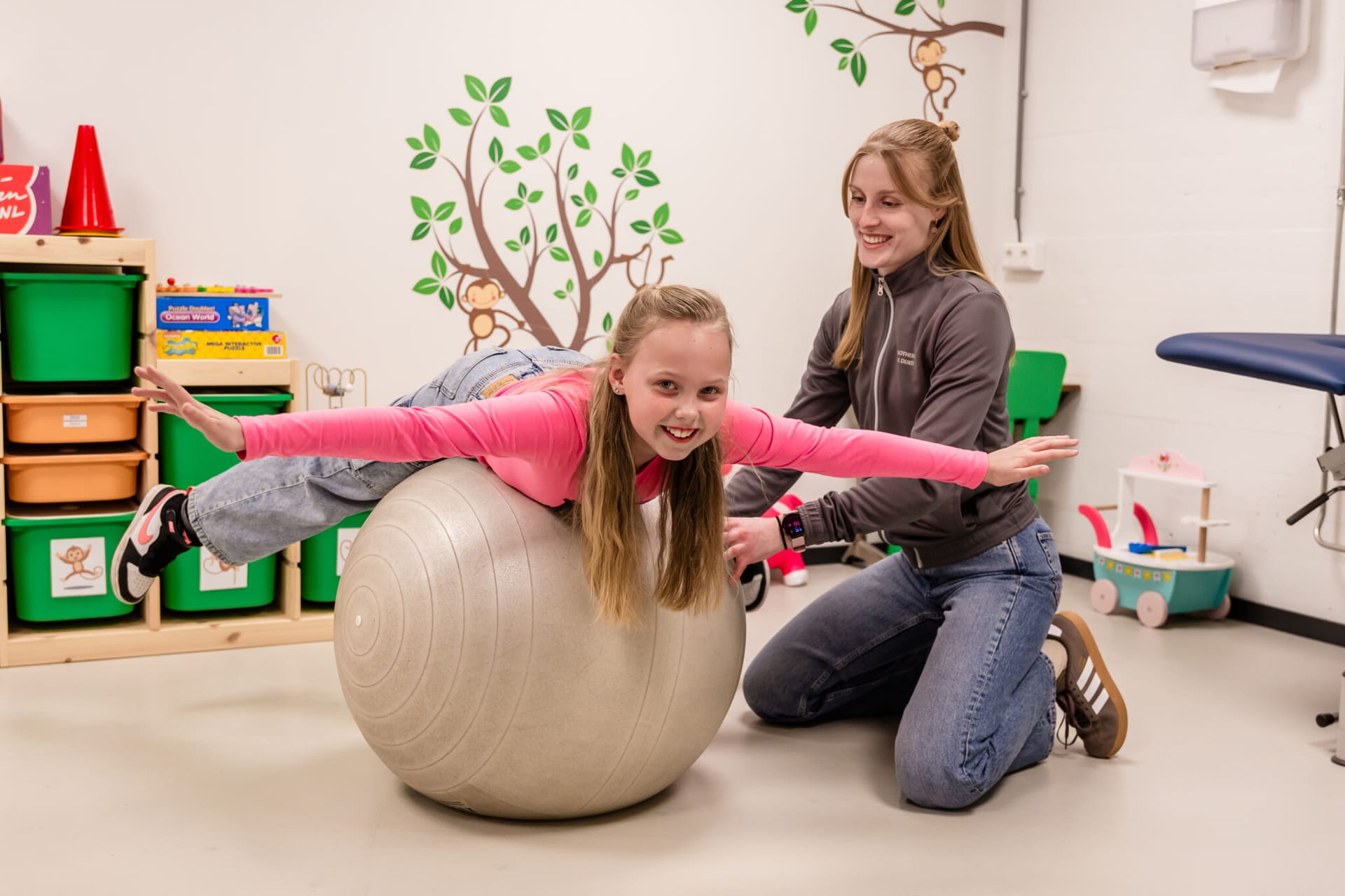 Een kinderfysiotherapeut begeleidt een meisje dat met gestrekte armen op een grote oefenbal balanceert in de oefenruimte.
