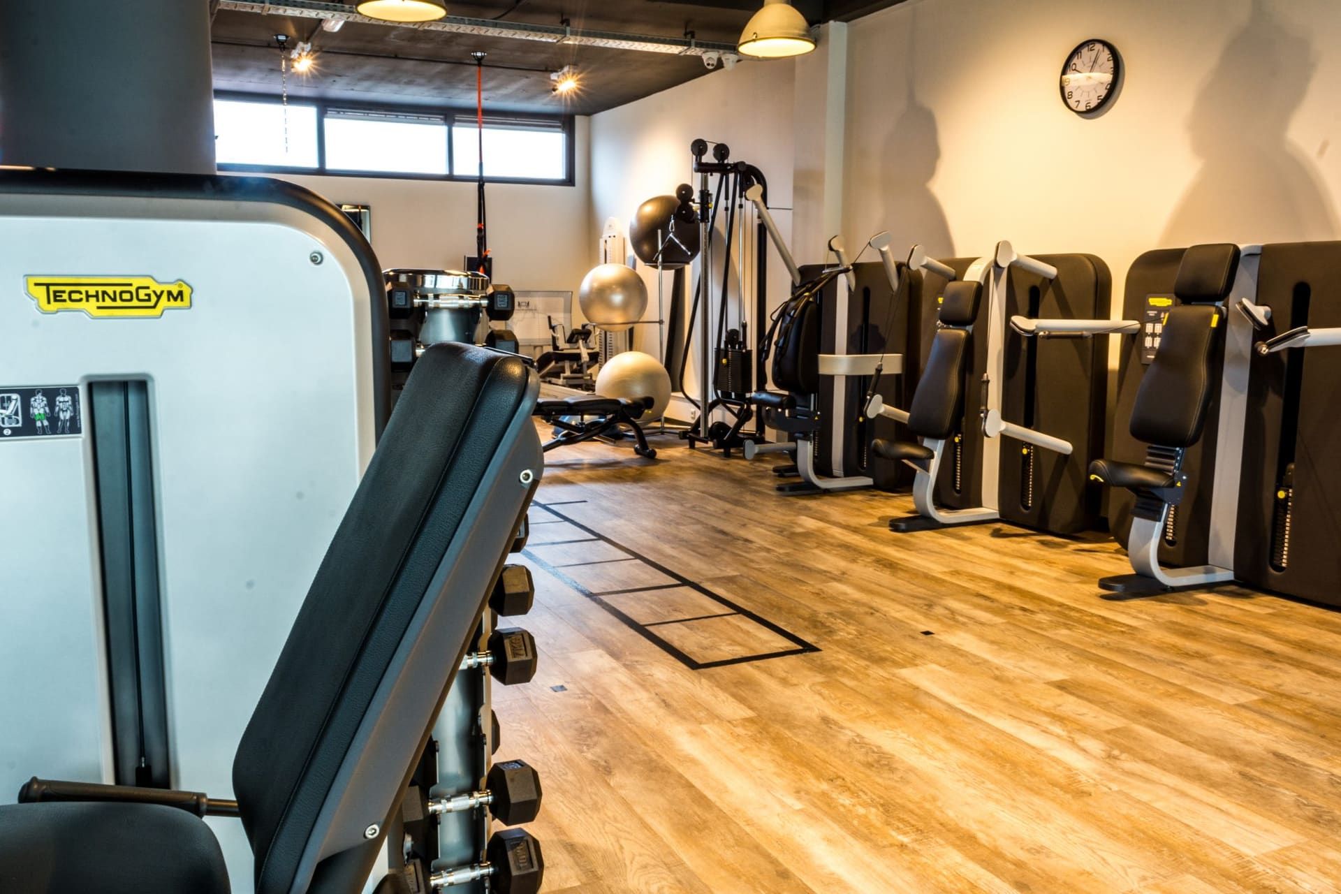 De oefenzaal met Technogym krachtapparaten langs de wand en een houten vloer.
